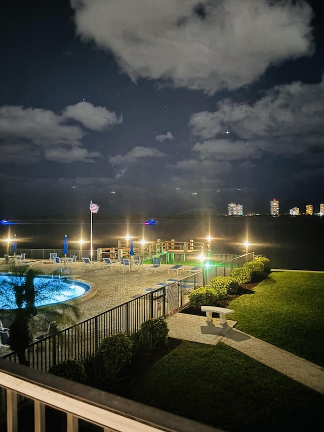 pool at night featuring a lawn, a water view, and a patio