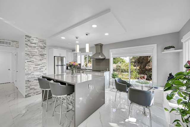 kitchen featuring white cabinets, wall chimney exhaust hood, stainless steel fridge, decorative light fixtures, and a kitchen island