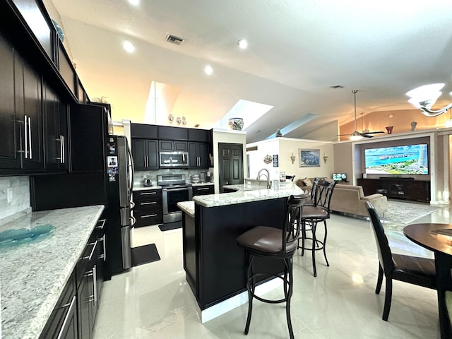 kitchen featuring backsplash, a center island with sink, vaulted ceiling with skylight, light stone countertops, and appliances with stainless steel finishes