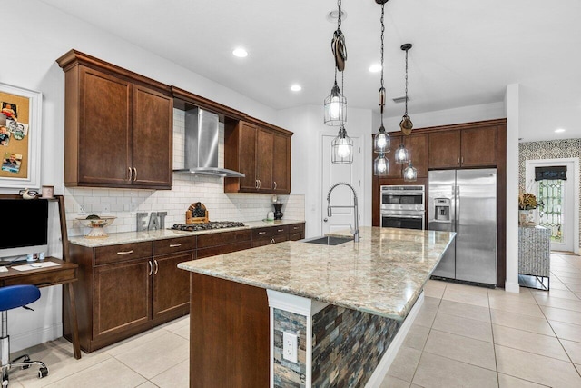 kitchen featuring sink, wall chimney range hood, appliances with stainless steel finishes, a kitchen island with sink, and light stone countertops