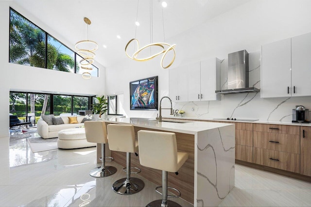 kitchen with white cabinetry, sink, wall chimney exhaust hood, pendant lighting, and a center island with sink