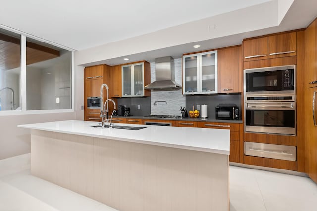 kitchen featuring sink, wall chimney range hood, an island with sink, stainless steel appliances, and backsplash