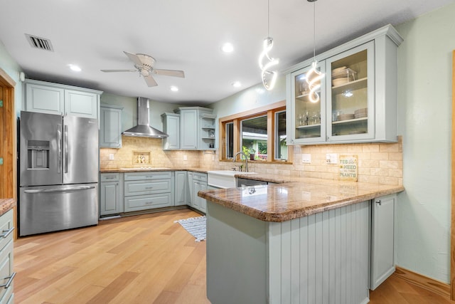 kitchen with stainless steel fridge, wall chimney exhaust hood, glass insert cabinets, a peninsula, and hanging light fixtures