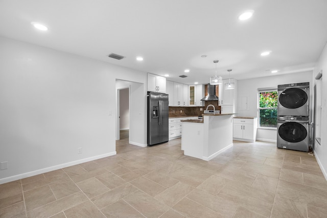 kitchen with stacked washer and dryer, stainless steel fridge, an island with sink, decorative light fixtures, and wall chimney range hood