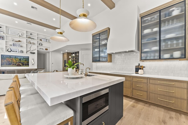 kitchen featuring decorative light fixtures, stainless steel microwave, beamed ceiling, a kitchen island with sink, and decorative backsplash