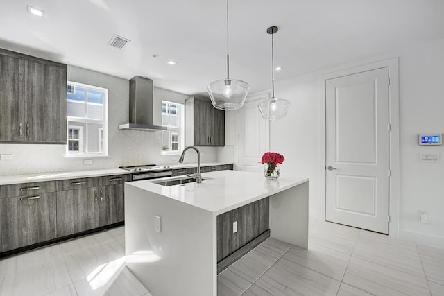 kitchen featuring decorative backsplash, sink, wall chimney range hood, hanging light fixtures, and an island with sink