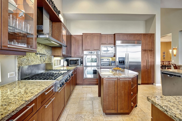 kitchen featuring light stone countertops, backsplash, wall chimney exhaust hood, built in appliances, and a kitchen island