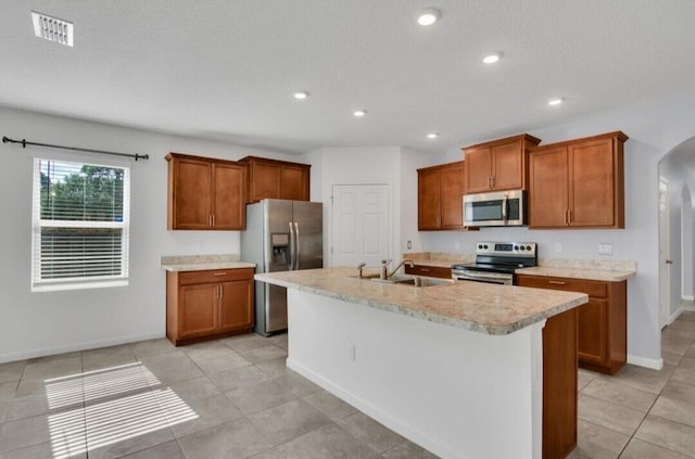 kitchen with a kitchen island with sink, sink, light tile patterned floors, a textured ceiling, and appliances with stainless steel finishes