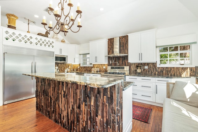 kitchen featuring wall chimney range hood, pendant lighting, lofted ceiling, a kitchen island with sink, and appliances with stainless steel finishes