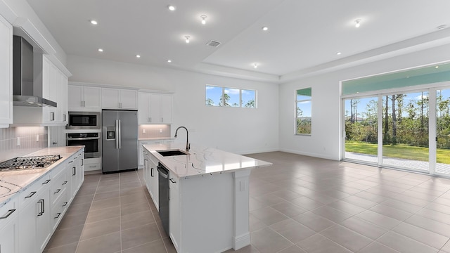 kitchen with stainless steel appliances, wall chimney range hood, sink, a center island with sink, and white cabinets