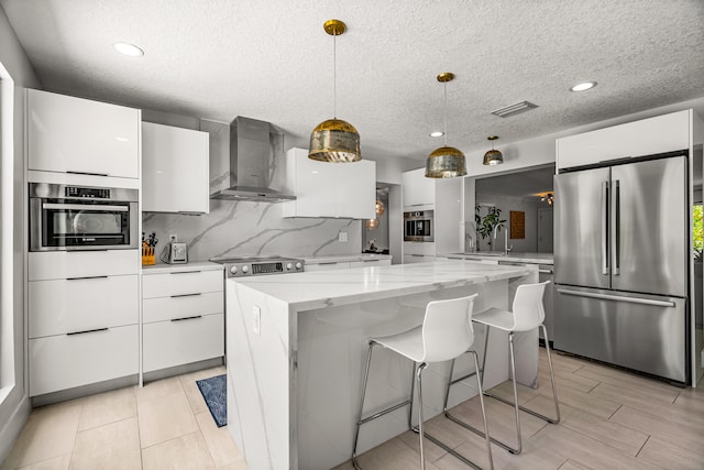 kitchen featuring appliances with stainless steel finishes, wall chimney exhaust hood, white cabinets, a kitchen island, and hanging light fixtures