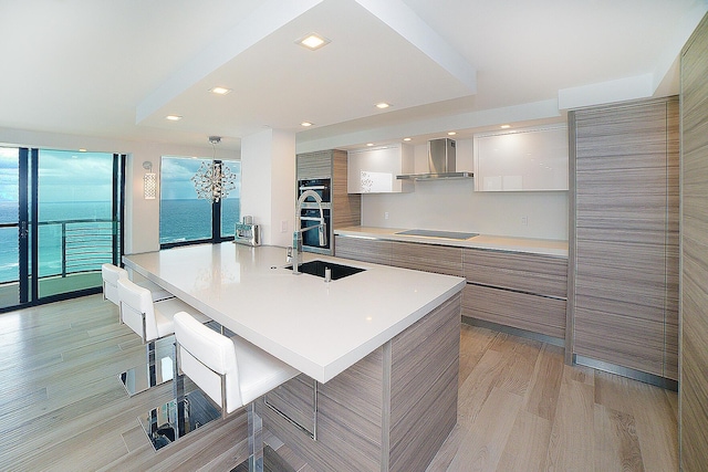 kitchen with wall chimney exhaust hood, a kitchen bar, black electric stovetop, a water view, and light wood-type flooring