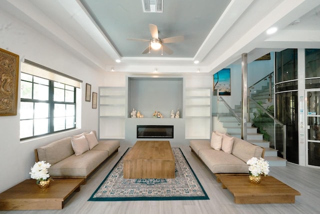living room with visible vents, a glass covered fireplace, a raised ceiling, ceiling fan, and stairs