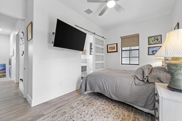 bedroom featuring light wood finished floors, a barn door, visible vents, baseboards, and a ceiling fan