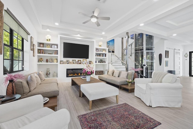 living area featuring a tray ceiling, a barn door, a glass covered fireplace, ceiling fan, and stairs