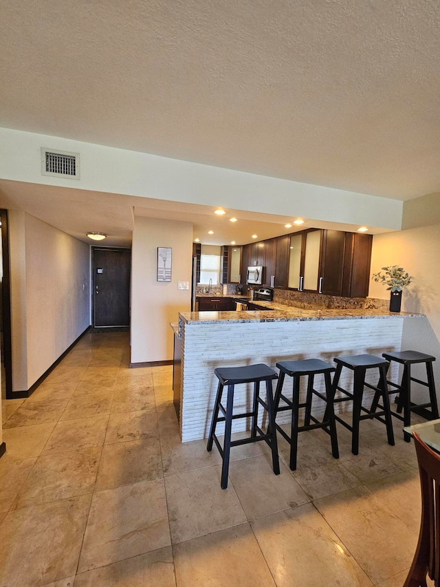 kitchen with a breakfast bar, kitchen peninsula, a textured ceiling, light stone counters, and dark brown cabinetry