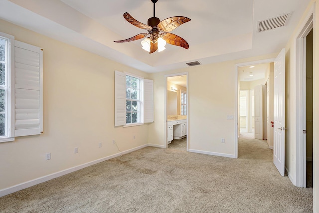 unfurnished bedroom with a tray ceiling, connected bathroom, ceiling fan, and light carpet