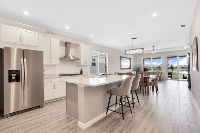 kitchen with white cabinetry, wall chimney range hood, light hardwood / wood-style flooring, an island with sink, and appliances with stainless steel finishes