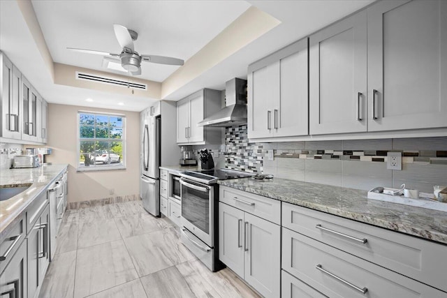 kitchen featuring light stone counters, a ceiling fan, stainless steel appliances, wall chimney exhaust hood, and tasteful backsplash