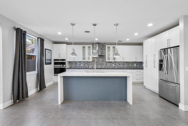 kitchen featuring stainless steel appliances, wall chimney range hood, white cabinetry, hanging light fixtures, and an island with sink