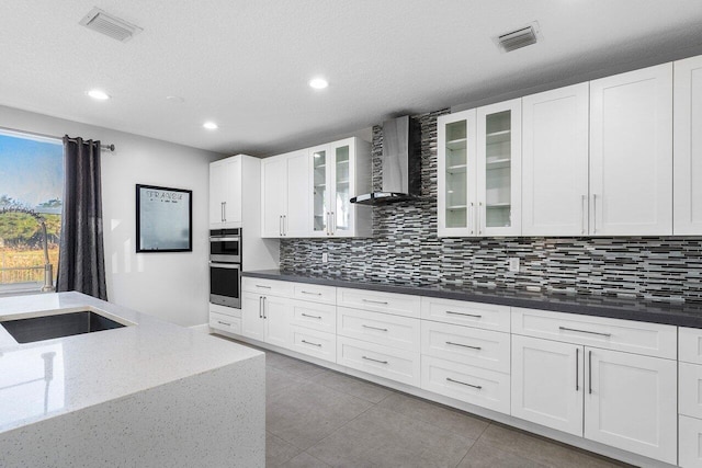 kitchen featuring black electric stovetop, double oven, wall chimney range hood, dark stone countertops, and white cabinets