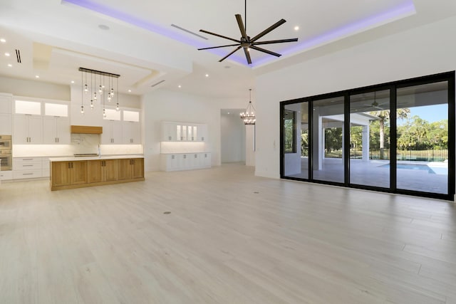 unfurnished living room with ceiling fan with notable chandelier, a tray ceiling, a towering ceiling, and light wood-type flooring