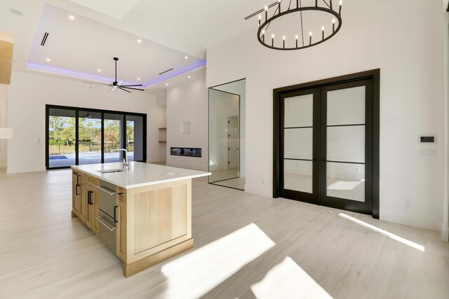 kitchen featuring a raised ceiling, light brown cabinets, a kitchen island with sink, ceiling fan with notable chandelier, and sink