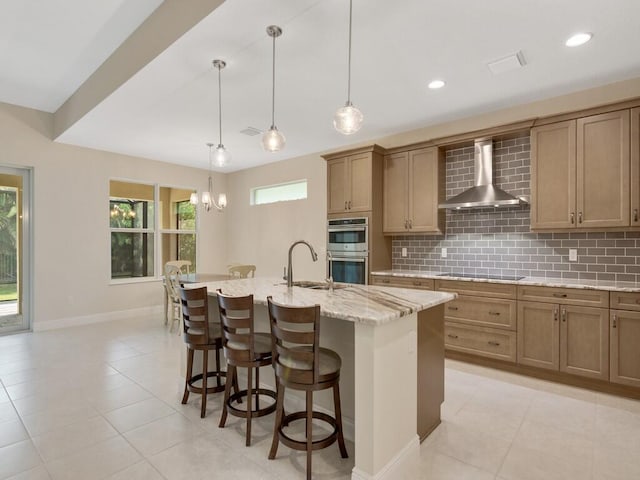 kitchen featuring black electric stovetop, wall chimney range hood, sink, an island with sink, and double oven