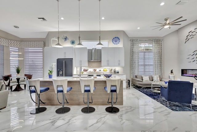 kitchen featuring white cabinetry, wall chimney range hood, stainless steel fridge with ice dispenser, decorative light fixtures, and a breakfast bar