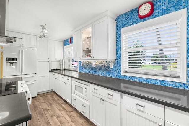 kitchen with white appliances, dark countertops, glass insert cabinets, ventilation hood, and white cabinetry