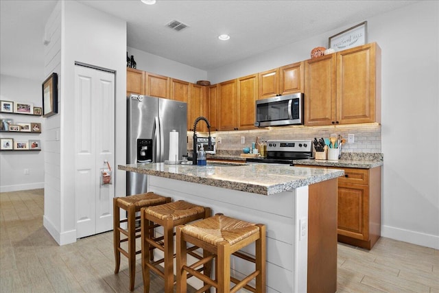 kitchen with a kitchen bar, a center island with sink, tasteful backsplash, light stone counters, and stainless steel appliances