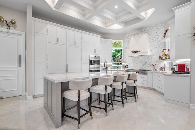 kitchen with white cabinetry, coffered ceiling, premium range hood, an island with sink, and a kitchen bar