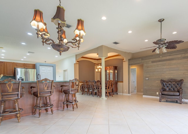 kitchen featuring stainless steel fridge with ice dispenser, decorative columns, a breakfast bar area, light tile patterned floors, and ceiling fan with notable chandelier