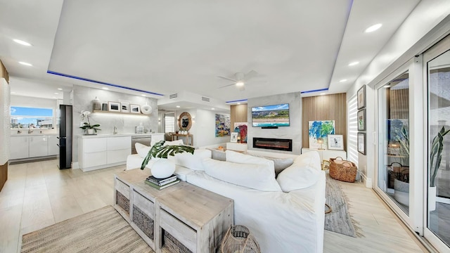 living room with a large fireplace, a tray ceiling, ceiling fan, sink, and light hardwood / wood-style floors