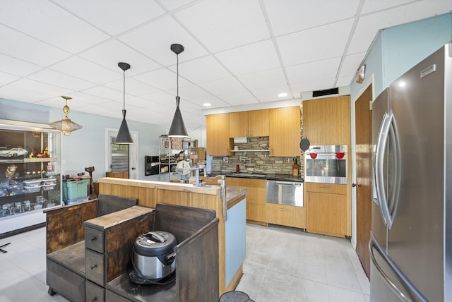 kitchen with decorative backsplash, appliances with stainless steel finishes, a drop ceiling, a center island with sink, and hanging light fixtures