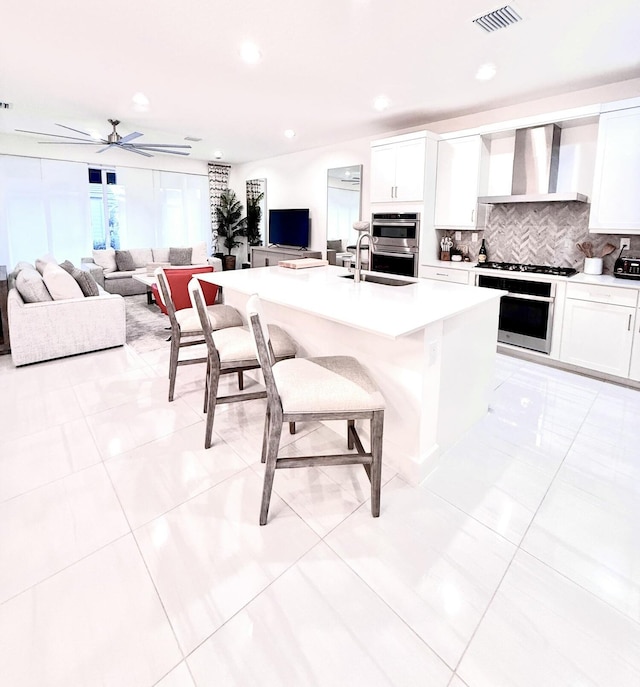 kitchen featuring tasteful backsplash, wall chimney exhaust hood, light tile patterned floors, a center island with sink, and white cabinetry