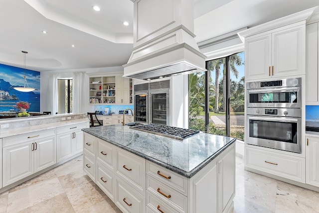 kitchen featuring a kitchen island with sink, stainless steel appliances, light stone countertops, premium range hood, and pendant lighting