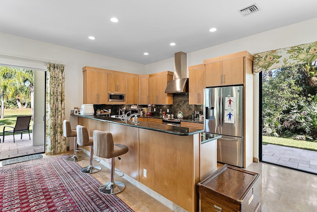 kitchen with appliances with stainless steel finishes, kitchen peninsula, dark stone counters, wall chimney exhaust hood, and tasteful backsplash