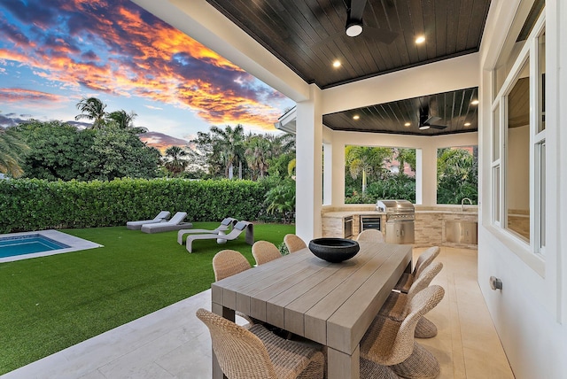 patio terrace at dusk with a grill, ceiling fan, a lawn, and exterior kitchen