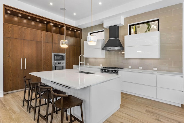 kitchen featuring stainless steel appliances, wall chimney exhaust hood, an island with sink, and light hardwood / wood-style floors