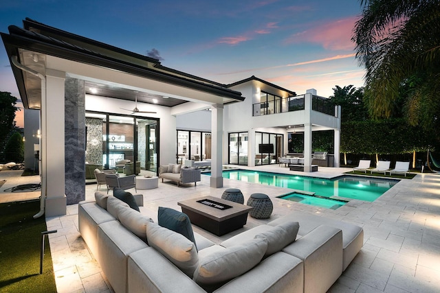 back house at dusk with ceiling fan, a pool with hot tub, a balcony, an outdoor living space with a fire pit, and a patio area