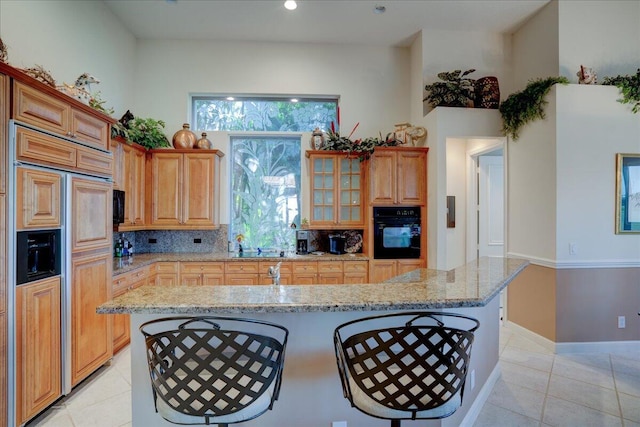 kitchen with light stone countertops, backsplash, an island with sink, black oven, and paneled built in fridge