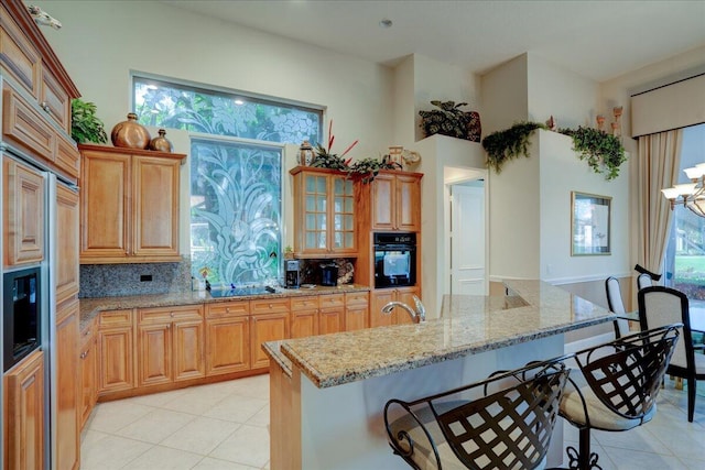 kitchen with decorative backsplash, light stone countertops, a center island with sink, black oven, and a breakfast bar area