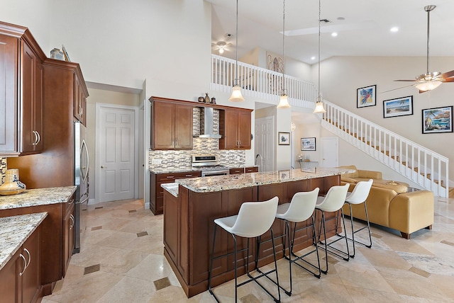 kitchen featuring high vaulted ceiling, wall chimney exhaust hood, an island with sink, appliances with stainless steel finishes, and decorative light fixtures