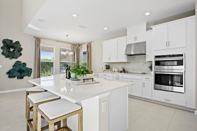 kitchen with double oven, a kitchen island with sink, white cabinetry, and pendant lighting