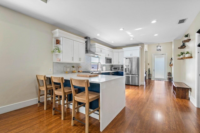 kitchen with light countertops, appliances with stainless steel finishes, white cabinets, wall chimney range hood, and a peninsula