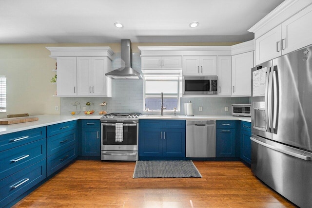 kitchen with stainless steel appliances, wall chimney range hood, blue cabinetry, and light countertops