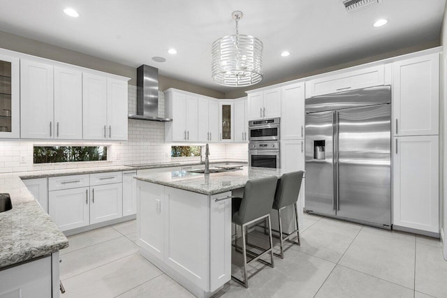 kitchen with a kitchen island with sink, white cabinets, wall chimney range hood, hanging light fixtures, and stainless steel appliances