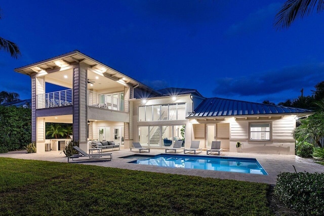 back house at night with an outdoor hangout area, a balcony, a patio, ceiling fan, and a yard