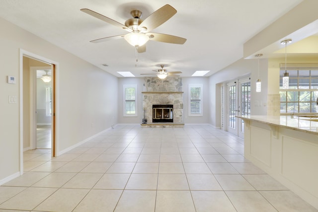 unfurnished living room with a skylight, a stone fireplace, ceiling fan, and light tile patterned flooring